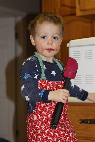 Child in apron with spoon in hand and baking mixture on face.